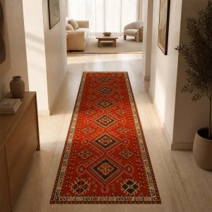 فرشینه کناره طرح عشایری هزارلوز با زمینه قرمز و نقوش هندسی، پهن شده روی کفپوش چوبی در یک راهروی مدرن و نورگیر که به اتاق نشیمنی با مبلمان مینیمال و رنگ‌های خنثی منتهی می‌شود.